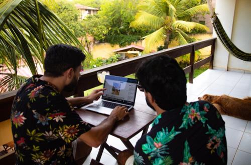two men sitting at a table with a laptop on a balcony at Iduna Hostel in Pipa