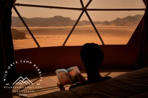 une personne assise dans un lit donnant par une fenêtre sur le désert dans l'établissement Wild Oryx Camp Bubbles, à Wadi Rum
