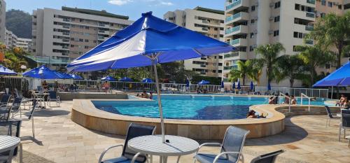 a pool with tables and chairs and blue umbrellas at Apartamento por temporada RJ próximo ao Riocentro, Parque Olímpico e Jeunesse Arena in Rio de Janeiro