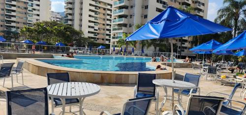 a swimming pool with tables and chairs and blue umbrellas at Apartamento por temporada RJ próximo ao Riocentro, Parque Olímpico e Jeunesse Arena in Rio de Janeiro