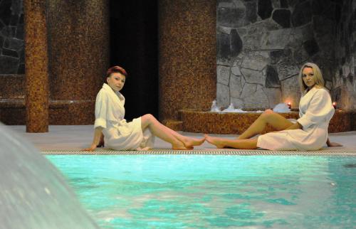 two women in white robes sitting next to a swimming pool at Hotel Karino Spa in Polańczyk