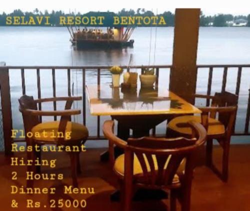 a wooden table and chairs with a boat in the water at Selavi Resort Bentota in Bentota