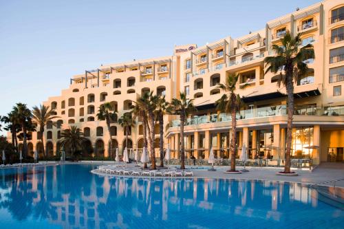 un grand bâtiment avec des palmiers et une piscine dans l'établissement Hilton Malta, à San Ġiljan