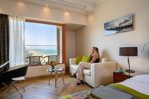a woman sitting on a couch in a room with a large window at Carlton Tel Aviv Hotel – Luxury on the Beach in Tel Aviv
