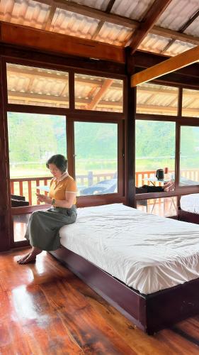a woman sitting on a bed in a room with windows at Đồng Lâm Ecolodge Homestay in Hữu Lũng