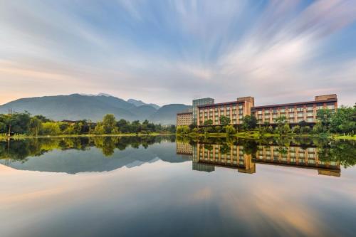 Le Méridien Emei Mountain Resort في إيمايشان: مبنى بجانب بحيرة فيها جبال في الخلفية