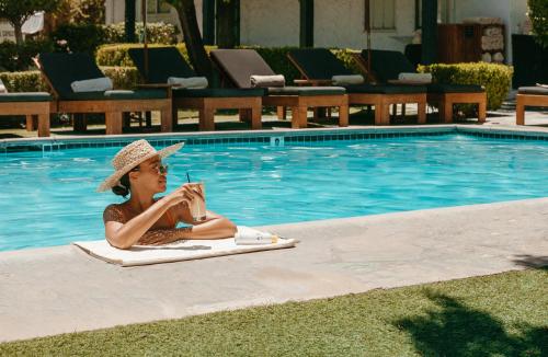 Una donna con un cappello seduta su un asciugamano in piscina di Avalon Hotel & Bungalows Palm Springs, a Member of Design Hotels a Palm Springs