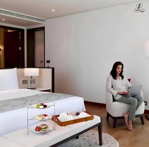 a woman sitting in a chair next to a bed with a laptop at The Leela Bhartiya City Bengaluru in Bengaluru
