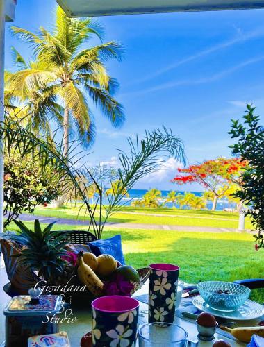 a table with food and a view of a palm tree at Accès Direct Plage, Vue Mer d'exception, Les Gwada Studios, Village Vacances in Sainte-Anne