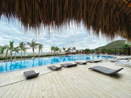 uma piscina com espreguiçadeiras e um guarda-sol de palha em Sensacional apartamento en Samaria Club de Playa em Santa Marta