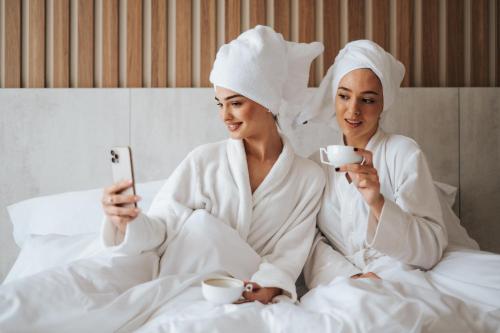 two women sitting in bed looking at a cell phone and drinking coffee at Hotel Kod Barića in Grude