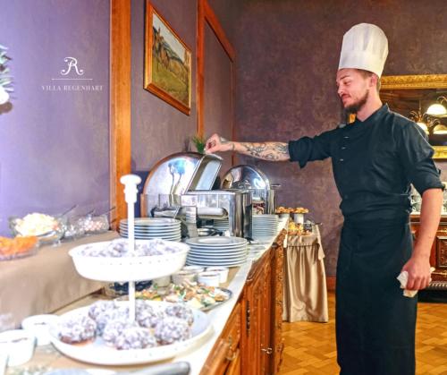 Un hombre con un gorro de chef de pie frente a una mesa con comida. en Wellness & Spa hotel Villa Regenhart, en Jeseník