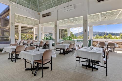 un restaurante con mesas y sillas blancas y ventanas grandes en Sanctuary Resort Motor Inn en Coffs Harbour