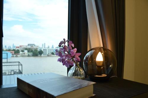 a book and a vase of flowers on a table with a window at NORN Riverside Bangkok Hotel - นอนริมน้ำ in Bangkok