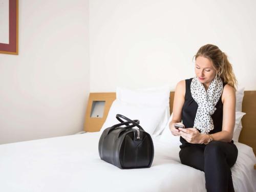 a woman sitting on a bed looking at her cell phone at Novotel Paris Rueil Malmaison in Rueil-Malmaison