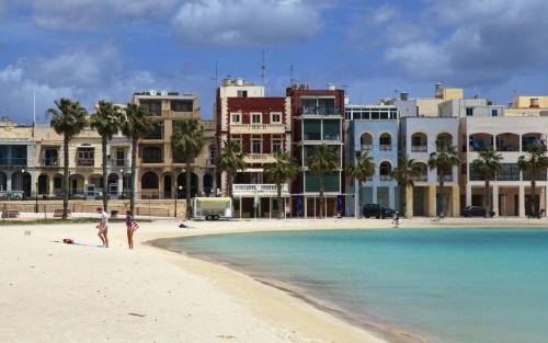 a group of people walking on a beach with buildings at A day at Dee's in Birżebbuġa