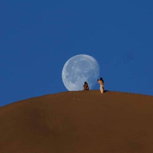 two people sitting on top of a sand dune with the moon at berber sahara in Zagora