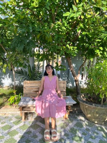 a woman in a pink dress sitting on a bench under a tree at Laoxao Homestay in Hoi An