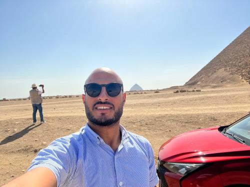 a man standing next to a red car in the pyramids at Sahure Pyramid View Inn in Cairo