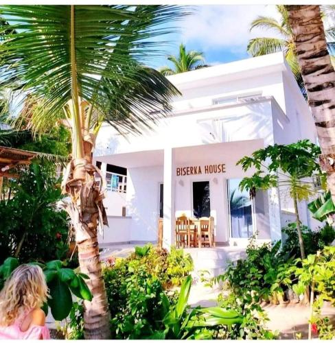 a woman sitting in front of a white house with a palm tree at Sipano Beach Lodge Kiwengwa in Kiwengwa
