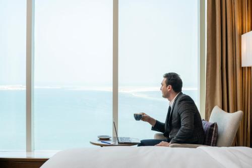 um homem sentado numa cama com um portátil e uma chávena de café em Delta Hotels by Marriott City Center Doha em Doha