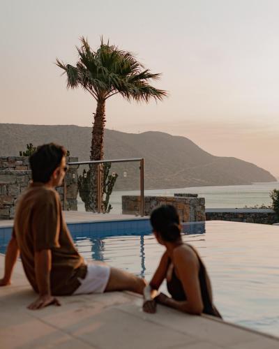 a man and a woman sitting next to a swimming pool at Phāea Blue - Small Luxury Hotels of the World in Elounda
