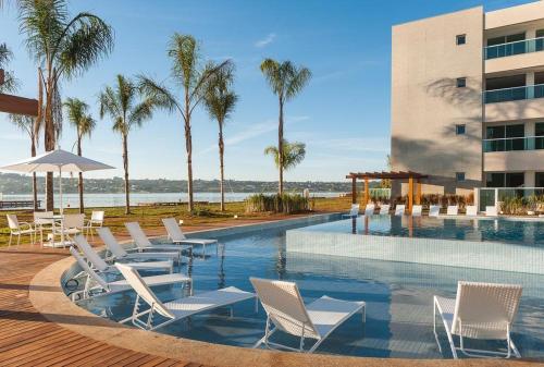 a pool with white chairs and a building and palm trees at Luxuoso Flat Brisas do Lago Terraço Aconchegante in Brasília
