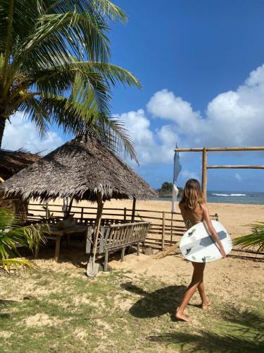 Pacific Surf and Yoga في Baras: فتاة صغيرة تمشي على الشاطئ مع لوح ركوب الأمواج