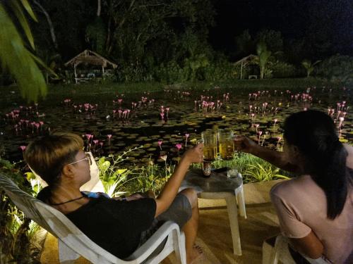 zwei Personen sitzen an einem Tisch mit Getränken vor einem Teich in der Unterkunft Bua Daeng Homestay Resort in Amphoe Kumphawapi