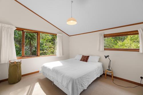 a white bedroom with a bed and two windows at Treetop Sanctuary - Titirangi Holiday Home in Auckland