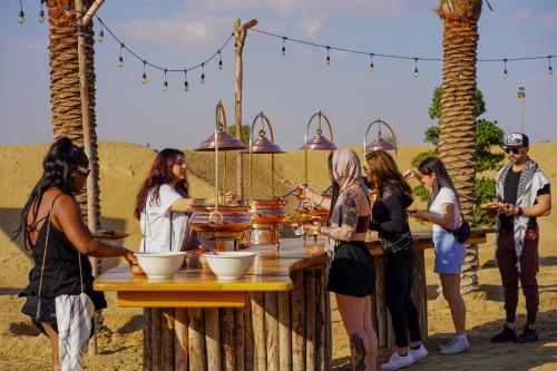 eine Gruppe von Menschen, die um einen Tisch mit Essen stehen in der Unterkunft Al Marmoom Oasis “Luxury Camping & Bedouin Experience” in Dubai