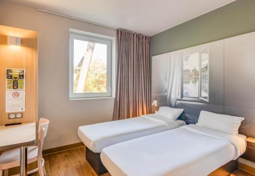 two beds in a room with a table and two windows at B&B HOTEL Bordeaux Langon in Langon