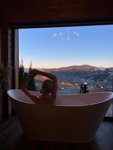 a woman in a bath tub in a room with a window at Under Sky Slavske - Котеджі з Теплим басейном ,чаном-джакузі, панорамною ванною та PlayStation 5 in Slavske