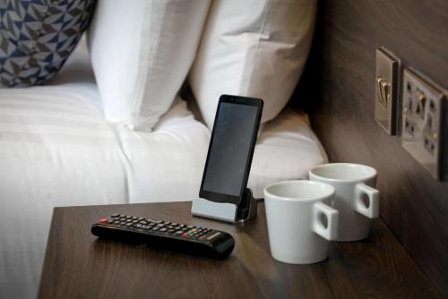 a remote control and two cups on a table in front of a bed at Trafford Hall Hotel Manchester, Trademark Collection by Wyndham in Manchester