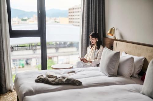京都市にあるザ ロイヤルパーク キャンバス 京都二条の窓を見ながらベッドに腰掛けている女性