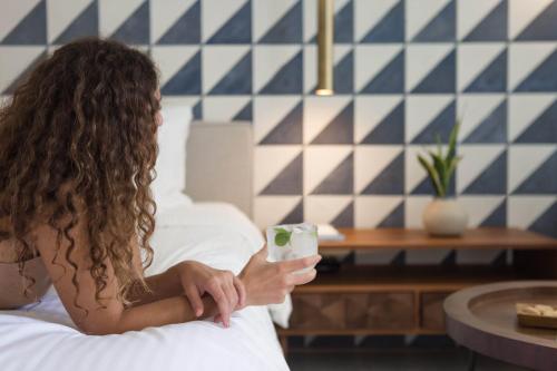 a woman sitting on a bed holding a glass of milk at Phāea Cretan Malia, a Member of Design Hotels in Malia