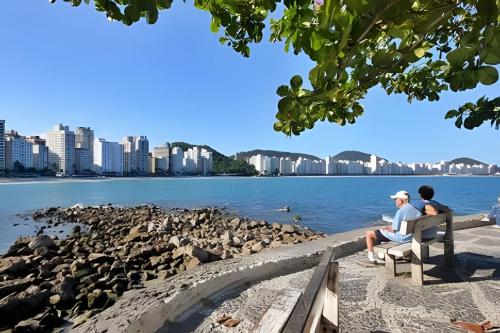 deux personnes assises sur un banc près de l'eau dans l'établissement Apartamento em Guarujá a 200 m da Asturias e Tombo, à Guarujá