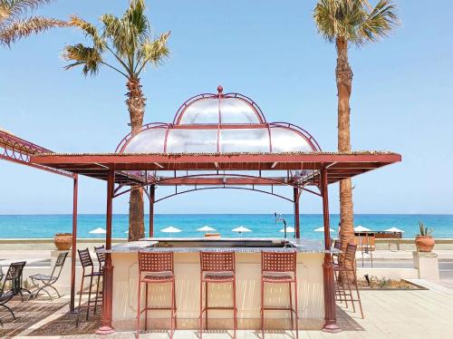 a bar with chairs and palm trees on the beach at Grecotel Plaza Beach House in Rethymno Town