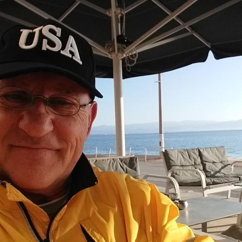 a man wearing a usa hat sitting under an umbrella at HELLAS PALACE ROOMS balcony in Sparti