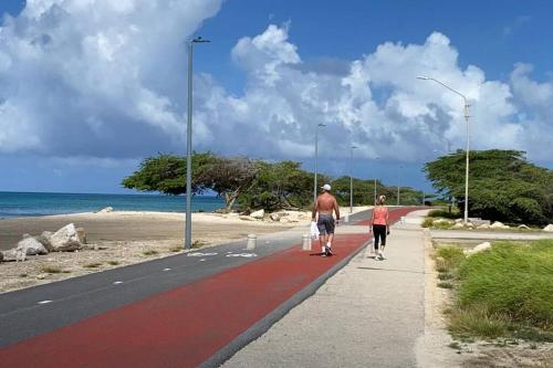 two people walking down a sidewalk next to the beach at Beautiful Vibes Sun Kiss Serenity your Stylish Apt Just Steps from Eagle Beach in Palm-Eagle Beach