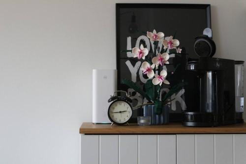 un reloj sentado en un estante con un jarrón de flores en Lille nest en Rødovre