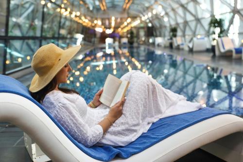 a woman sitting in a chair reading a book next to a swimming pool at Oryx Airport Hotel -Transit Only in Doha