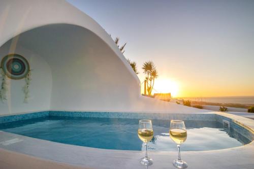 two glasses of wine next to a swimming pool with the sunset at Sun Angelos Oia - Luxury Cave Suites in Oia