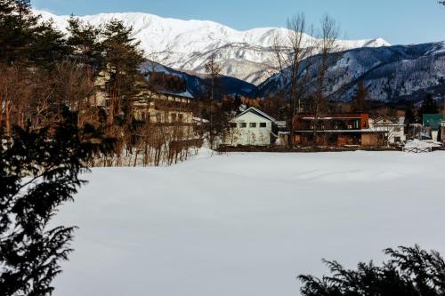 a snow covered field with a house and mountains at Hakuba Pension & Log Hotel Meteor in Hakuba