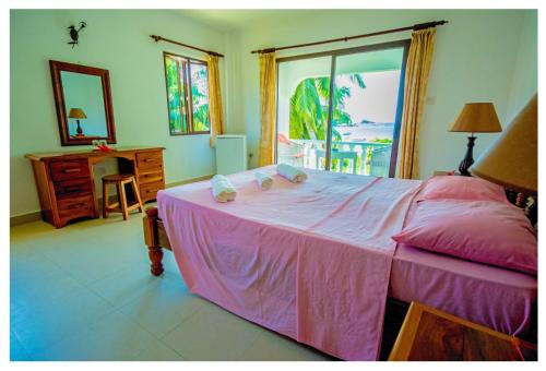 Schlafzimmer mit einem rosa Bett, einem Schreibtisch und einem Fenster in der Unterkunft Rosemary's Guesthouse in Praslin