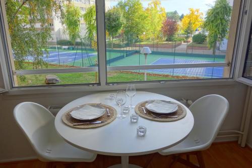 une table blanche avec deux chaises et une fenêtre avec un court de tennis dans l'établissement Arbres & Tennis, à Strasbourg