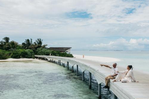 a couple sitting on a pier next to the beach at Cinnamon Hakuraa Huraa Maldives - All Inclusive in Meemu Atoll