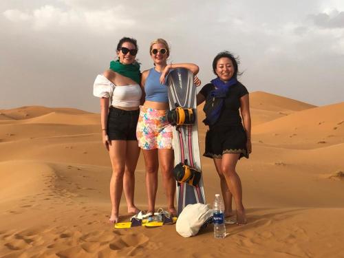 three women standing in the desert with a snowboard at Enjoy Moda Camp Merzouga tours- Camel sunset sunrise Quad Sunboarding ATV in Merzouga