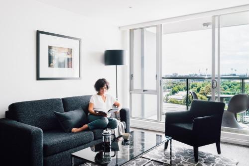 a woman sitting on a couch reading a book in a living room at Quay West Suites Melbourne in Melbourne