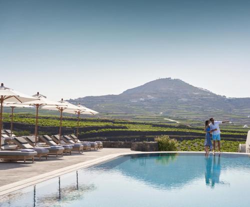 two people standing next to a swimming pool with a mountain at Vedema, a Luxury Collection Resort, Santorini in Megalochori